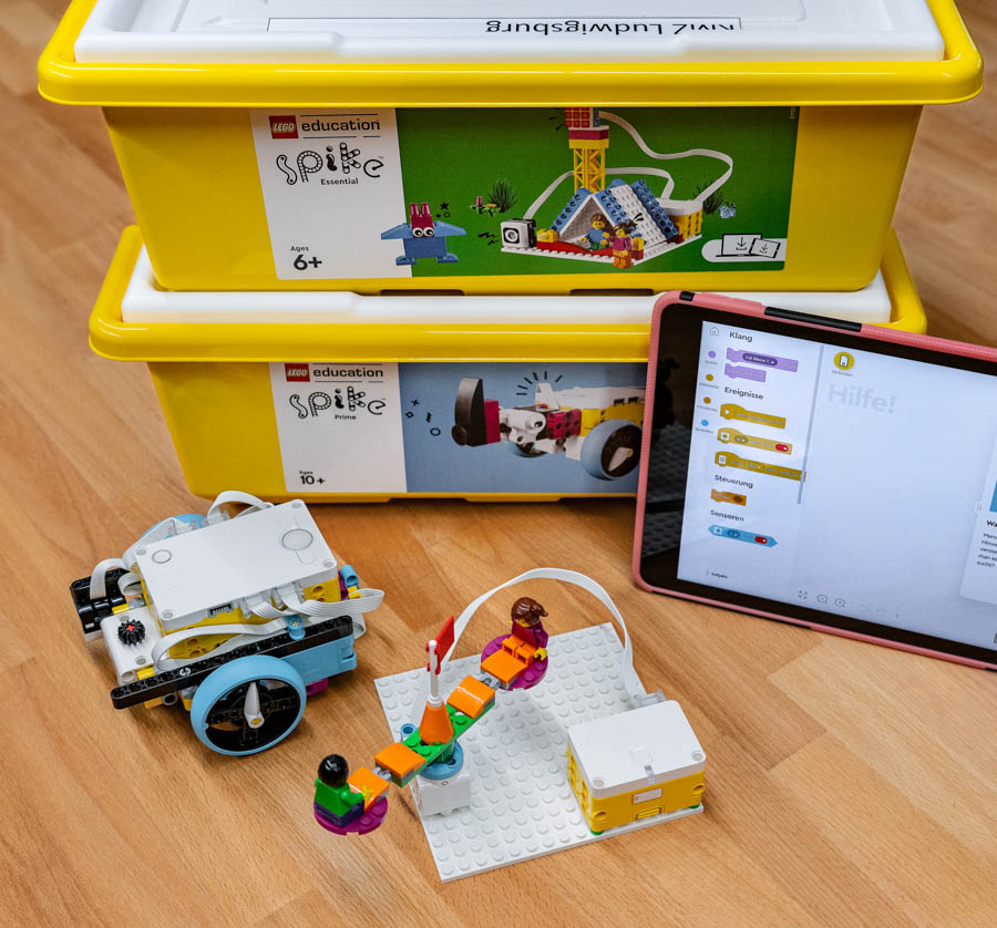 Dies ist ein Bild zeigt 2 Kisten Lego Spike Edukation. Vor der Kiste steht ein daraus gebautes Karussell das über einen kleinen Motor angetrieben wird. Ebenfalls steht dort ein iPad und ein kleiner MB Roboter